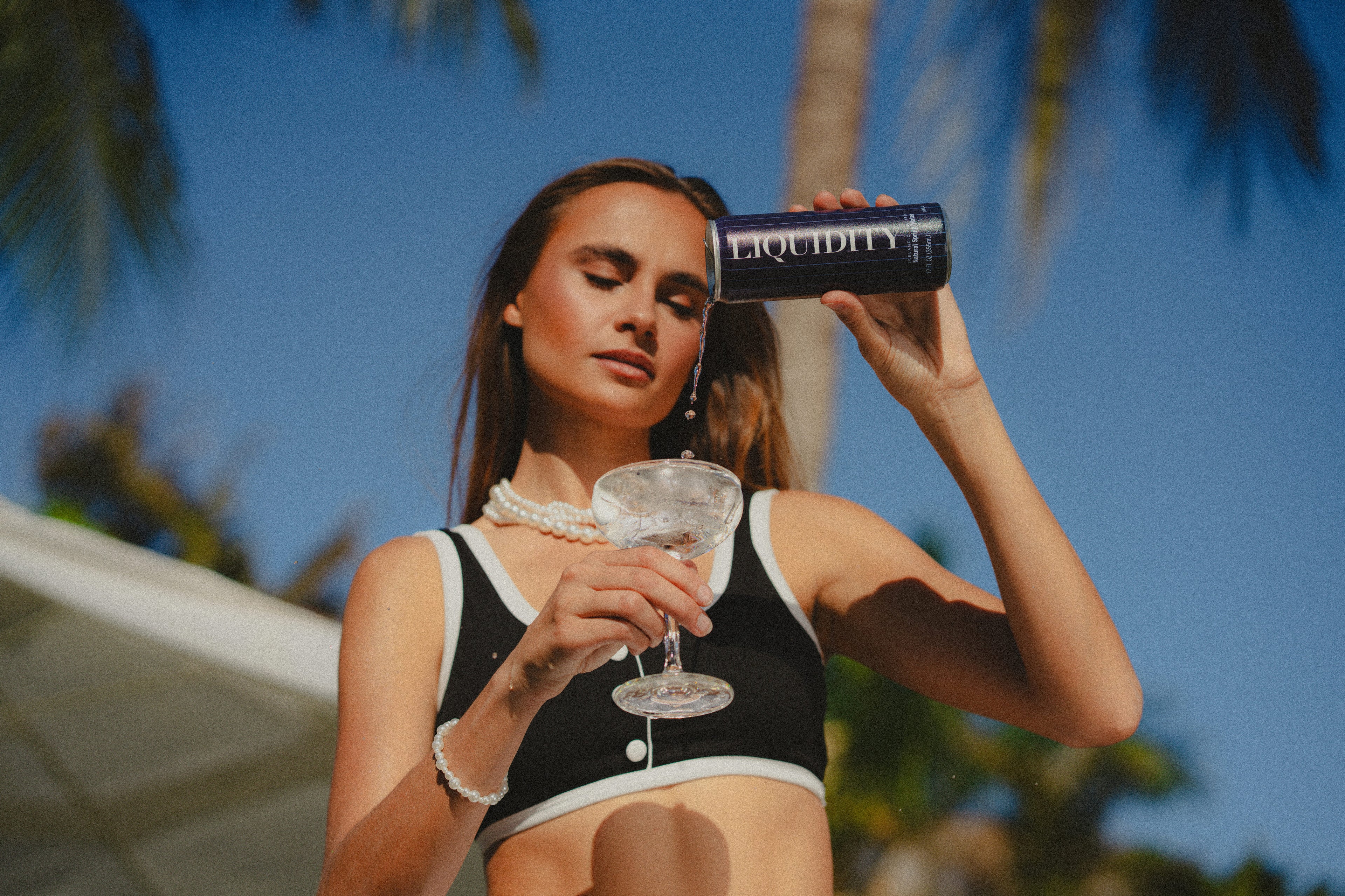 Woman in a black and white outfit holding a can of water and a glass against a blue sky with palm trees.